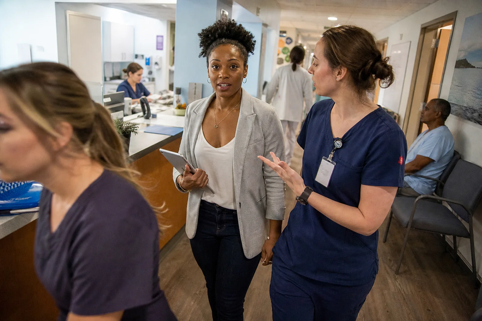 An administrator holding a tablet talking with a nurse in navy scrubs as they walk a clinic corridor, with the front desk and the waiting area behind them.