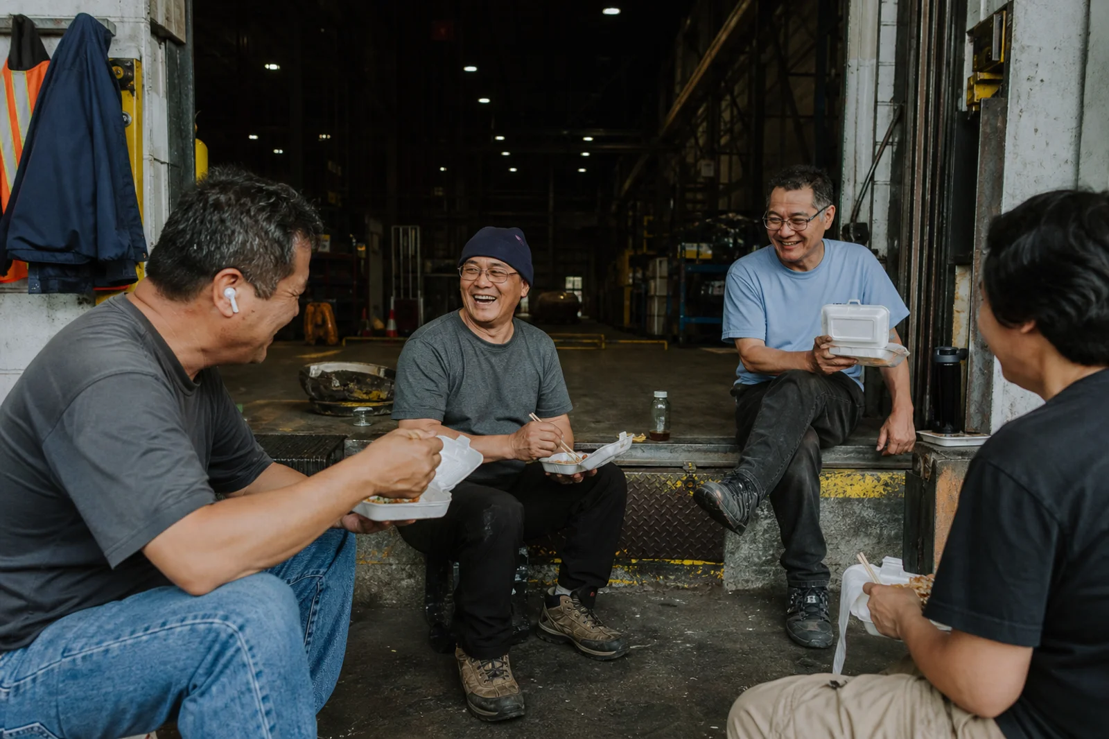 Four shop workers sitting on the edge of a loading dock at lunch, eating from takeout containers and laughing, with the plant dark behind them.